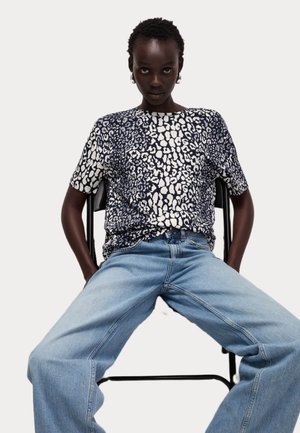 T-shirt à manches courtes en imprimé léopard bleu marine et blanc, avec une coupe ample, associé à un jean large bleu clair. Modèle assis sur une chaise noire.