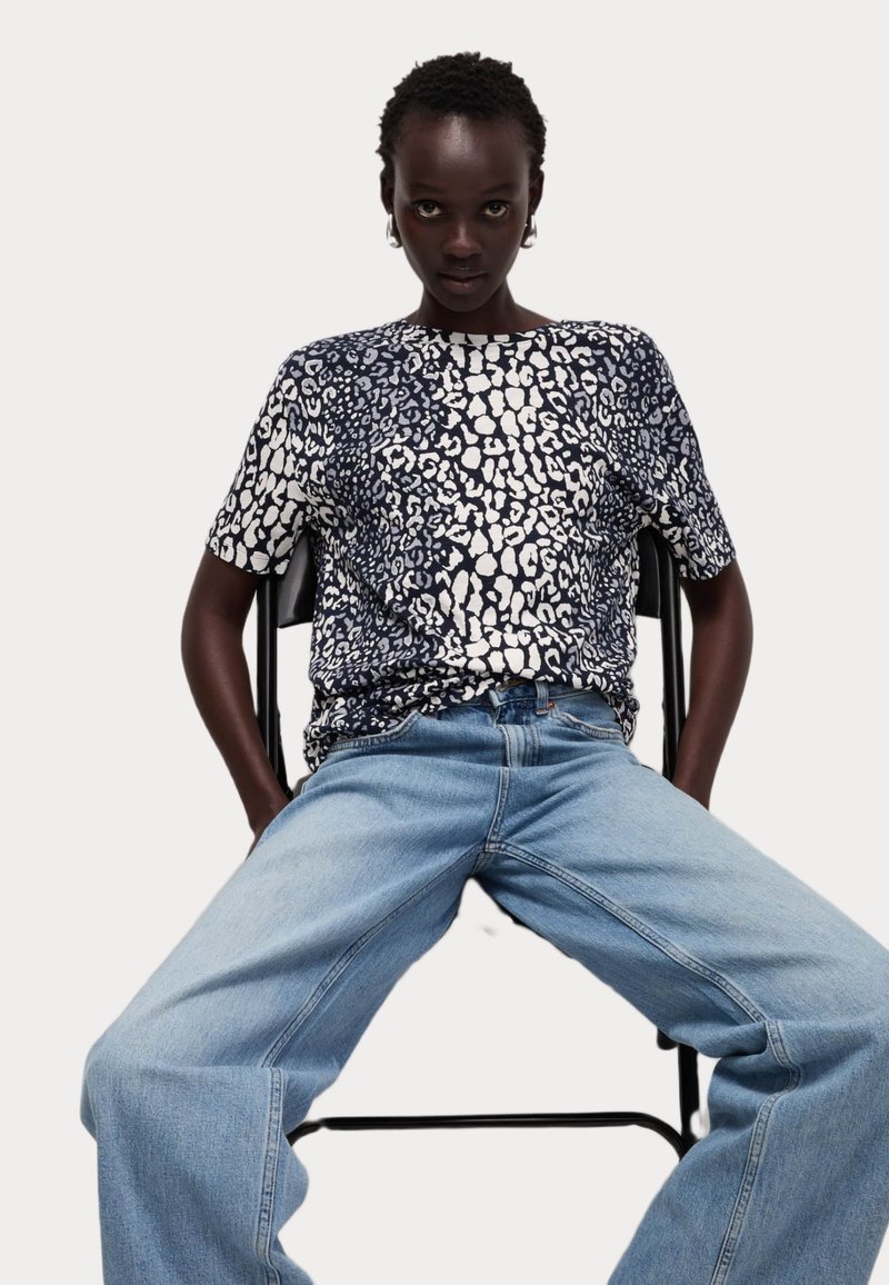 Tričko s krátkým rukávem v námořnické modré barvě s leopardím potiskem v bílé barvě a volným střihem, spárované s světle modrými širokými džíny. Model sedí na černé židli.
