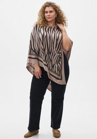 Poncho à motifs marron et noir avec un design fluide. Présente une bordure géométrique et une texture douce, associé à un pantalon noir et des chaussures marron.