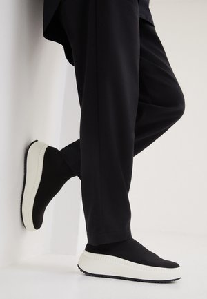 Person trägt schwarze Hose und schwarze Slip-on-Sneaker mit dicken weißen Sohlen, ein Fuß lehnt an einer weißen Wand.