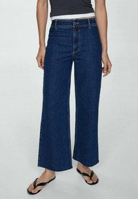 Jean denim bleu foncé à jambes larges avec une taille haute, doté d'une fermeture éclair et bouton à l'avant, et d'un léger effet délavé le long des jambes.