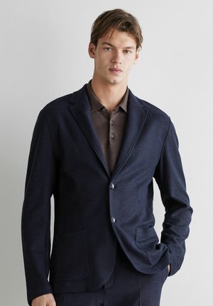 Junger Mann mit hellbraunen Haaren, der einen dunkelblauen Blazer über einem braunen Hemd mit Knöpfen trägt und vor einer schlichten hellen Wand steht.