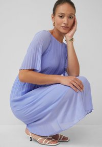 Hellblau-lila plissiertes Kleid mit kurzen, flatternden Ärmeln, kombiniert mit weißen Riemchen-Heels. Glatter Stoff mit subtiler Schattierung und fließendem Design.