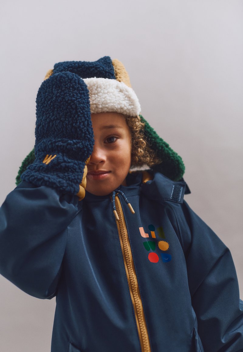 Enfant portant un manteau d'hiver bleu, un bonnet en polaire multicolore et des moufles assorties, couvrant un œil avec une moufle sur un fond uni.