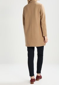 Cappotto di lana cammello con taglio dritto, maniche lunghe e spacco posteriore, abbinato a pantaloni scuri e scarpe in suede rosse. Dettaglio del colletto piatto.