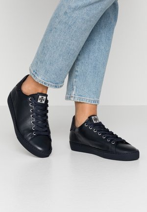 Osoba nosząca czarne skórzane sneakersy na sznurowadła i jasnoniebieskie skrócone dżinsy, stojąca na gładkiej białej podłodze na tle jednolitego tła.