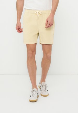 Mężczyzna w beżowych szortach z troczkami i białych tenisówkach z czarnymi paskami stoi na białym tle.