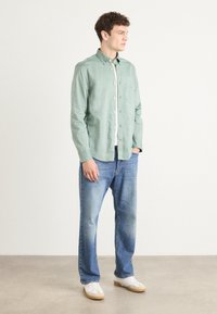 Lys grønn skjorte med knappestemming i et mykt stoff, kombinert med blå jeans og hvite joggesko. Skjorten har en brystlomme og klassisk krage.