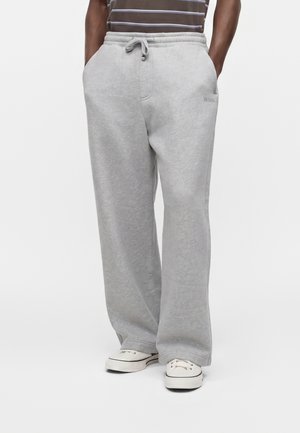 Personne portant un pantalon de jogging ample gris clair avec cordon et poches, assorti à des chaussures blanches décontractées, debout les mains dans les poches.