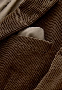 Tissu en velours côtelé marron avec des rainures verticales. Un mouchoir de poche beige partiellement visible dans une poche de poitrine, mettant en valeur la texture en couches.