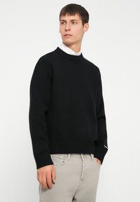 Fekete kötött pulóver ribbed nyakkal és mandzsettával. Fehér galléros ing fölé öltözve. Egyszerű dizájn, minta nélkül.