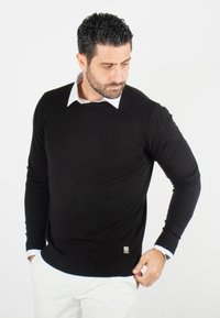 Pull en tricot noir avec un col rond, design ajusté, poignets et ourlet côtelés, superposé sur une chemise blanche à col. Petit logo en détail sur l'ourlet.