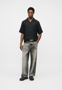 Άνδρας όρθιος, με το ένα χέρι στην τσέπη, φοράει μαύρο πουκάμισο κοντό μανίκι με κουμπιά, ξεθωριασμένο τζιν με φαρδιά μπατζάκια, μαύρη ζώνη και μαύρα παπούτσια.