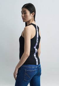 Γυναίκα που φοράει μαύρο αμάνικο τοπ με κάθετη λευκή ρίγα με το λογότυπο "EA7 Emporio" στην πλάτη, σε συνδυασμό με μπλε τζιν, στέκεται μπροστά σε απλό φόντο.