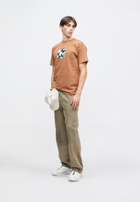 Brūns kokvilnas T-krekls ar grafisku attēlu krūtīs, kopā ar gaiši zaļām biksēm un baltām sporta kurpēm. Kreisajā rokā tiek turēta bēša cepure.