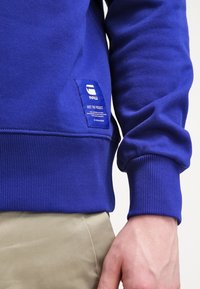 Blå sweatshirt i bomull med ribbade muddar, ett märke med logotyp och en mjuk textur. Fokuserar på komfort och avslappnad passform.