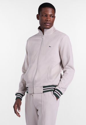 Mężczyzna w jasnobeżowej kurtce na zamek z ciemnozielonymi i białymi prążkowymi mankietami oraz paskiem w talii, ubrany w pasujące spodnie, stoi z rękami w kieszeniach.