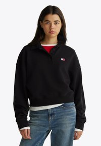Zwarte katoenen sweatshirt met een hoge kraag, voorzien van een knoopsluiting en een klein logo-embleem. G gedragen over een rode top en blauwe spijkerbroek.