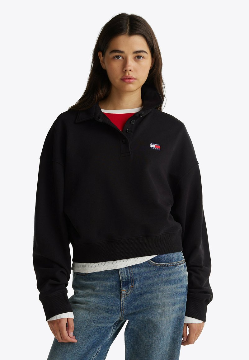 Zwarte katoenen sweatshirt met een hoge kraag, voorzien van een knoopsluiting en een klein logo-embleem. G gedragen over een rode top en blauwe spijkerbroek.