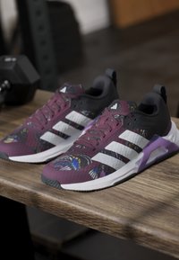 Ζευγάρι μωβ και μαύρα αθλητικά παπούτσια Adidas με λευκές ρίγες και πολύχρωμα μοτίβα πεταλούδας, τοποθετημένα σε ξύλινο παγκάκι κοντά σε γυμναστηριακό εξοπλισμό.