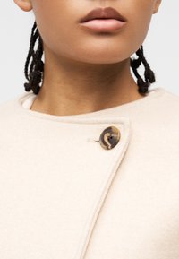 Κοντινό πλάνο ενός ατόμου που φοράει ένα μπεζ παλτό, στερεωμένο με ένα μεγάλο μάρμαρο κουμπί κοντά στον γιακά.