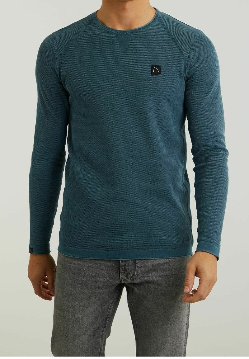 Teal långärmad skjorta i texturerat tyg, rund halsringning och svart logotypmärke. Grå jeans kompletterar outfiten.