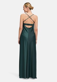 Langes, dunkelgrünes Satin-Kleid mit kreuzförmigem Rückendetail, das ein Rüschenelement über dem offenen Rücken und eine glatte, fließende Silhouette aufweist.