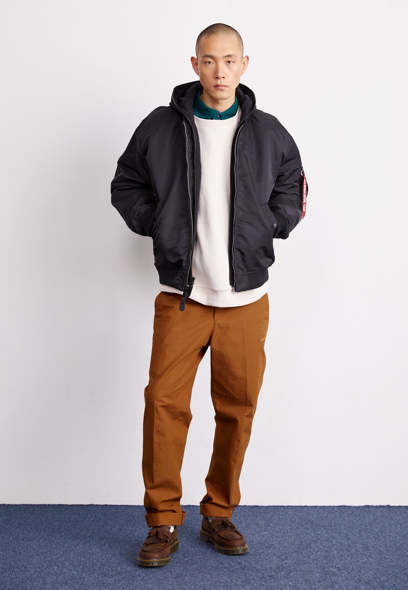 Veste bomber noire avec capuche, présentant une fermeture éclair et un détail de patch rouge. Portée par-dessus un sweat crème et associée à un pantalon et des chaussures bruns.