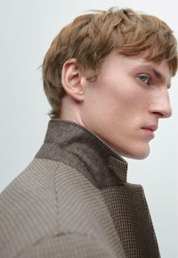 Brązowy płaszcz w jodełkę z wełnianym kołnierzem, o strukturalnym kroju i teksturowanej tkaninie. Zbliżenie profilu na neutralnym tle.