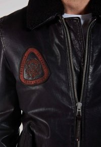 Gros plan sur une veste en cuir noir avec une fermeture éclair et un patch triangulaire rouge portant l'inscription "Chef d'équipe de la Patrouille de France" sur la poitrine.