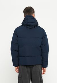 Mørkeblå pufferjakke med hætte, der har en quiltet tekstur og vandrette syninger. Bagsiden har et let cropped design.