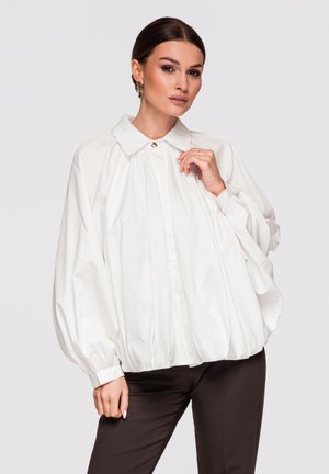 Kobieta ubrana w luźną, białą bluzkę z długim rękawem i złotym guzikiem przy kołnierzyku, w połączeniu z ciemnymi dopasowanymi spodniami, pozująca na jednolitym tle.