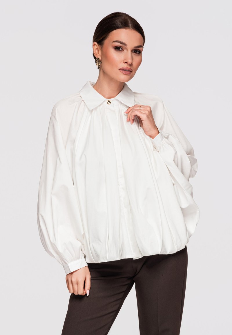 Femme portant un chemisier blanc ample à manches longues avec un bouton doré au col, associé à un pantalon foncé ajusté, posant devant un fond uni.