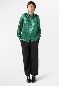 Groene metallic blouse met knoopsluiting aan de voorkant, gebogen zoom en lange mouwen, gecombineerd met zwarte wijde broek en enkellaarsjes.