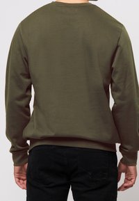 Olivi vihreä pitkähihainen svetari, joka on valmistettu pehmeästä kankaasta, pyöreällä kaula-aukolla ja ribbauslahkeilla, yhdistetty mustiin farkkuihin.