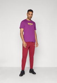 Lilla T-shirt med gul "BARÇA" tekst og Nike-logo, parret med røde sweatpants med en gul Nike-logo, båret med sorte træningssko.