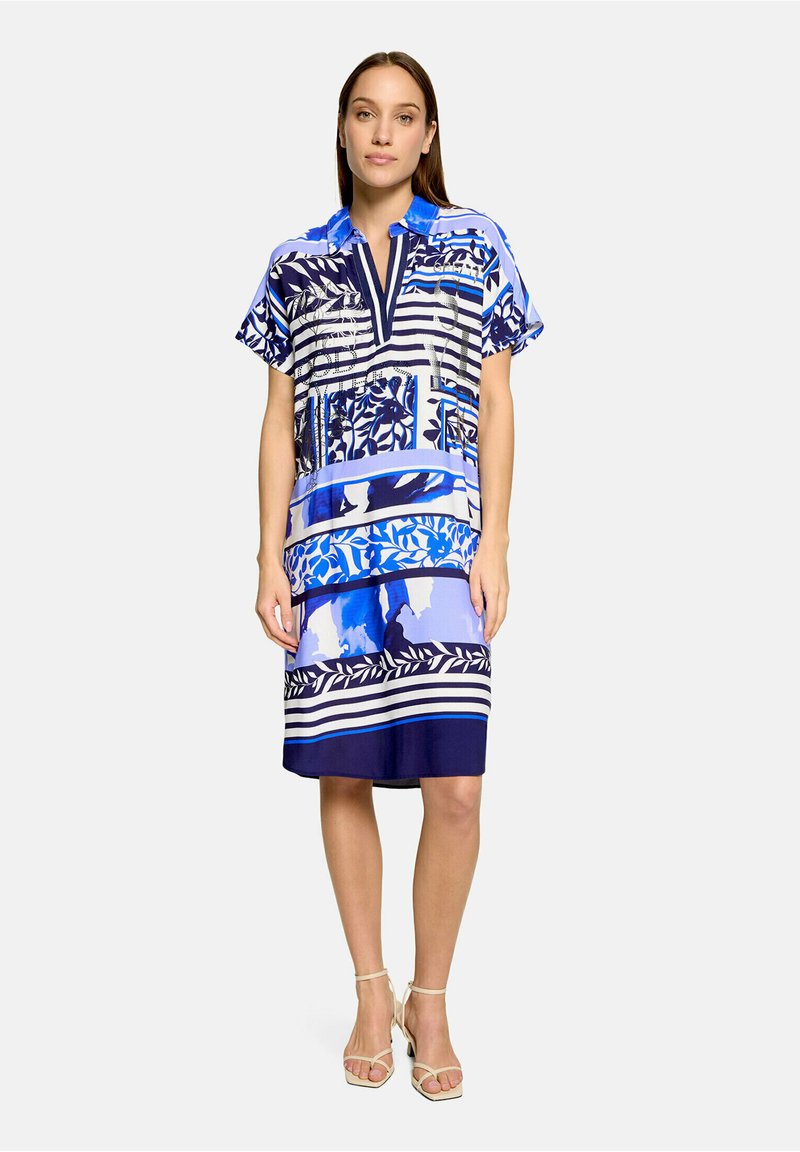 Femme portant une robe à manches courtes à motifs bleu, blanc et noir, longueur genou, avec col, encolure ouverte et sandales beiges à talons hauts, debout.