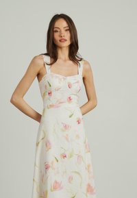 Weißes Blumenkleid aus leichtem Stoff, mit quadratischem Ausschnitt, breiten Trägern und einem zarten pinkfarbenen Blumenmuster im gesamten Kleid.