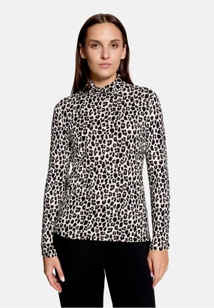 Vrouw draagt een hooggesloten, lange mouwen luipaardprint top en zwarte broek, staand tegen een effen lichte achtergrond.