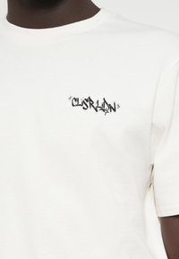 Λευκό βαμβακερό μπλουζάκι με στρογγυλή λαιμόκοψη, διακοσμημένο με ένα μαύρο λογότυπο σε στυλ γκράφιτι στο στήθος. Βραχίονες κοντοί, μαλακή υφή.