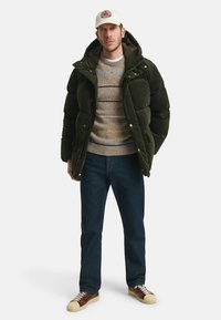 Grüne gepolsterte Jacke mit hohem Kragen, beige gestreiftes Sweatshirt, blaue Jeans und mehrfarbige Sneakers. Trägt eine weiße Mütze mit Logo.