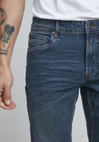 Temno modri kavbojki z ozkim fitom, ki imajo pet žepov, subtilno zbledelost in kovinske dele na pasu ter žepih.