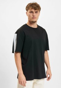 Juoda trumpomis rankovėmis marškinėliai su laisvu siluetu, apvalia iškirpte ir baltu plotu su grafika ant kairiojo rankovės.