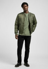 Oliwkowa bomberka z zapięciem na zamek i ściągaczami, połączona z dopasowaną zieloną koszulą, czarnymi spodniami i czarnymi skórzanymi butami.