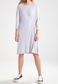 Vaaleanharmaa mekko, jossa on kietaisusuunnittelu, kolmivartiset hihat, ryppytyskohtia sivuilla ja virtaava siluetti. Yhdistetty musta-valkoisiin slip-on-kenkiä.