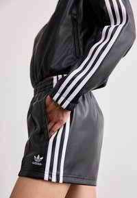 Zwart faux leren sportset met een ritsjack en shorts, accenten van witte drie-streep details en een geborduurd logo.