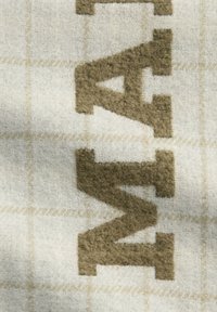 Geweven deken in beige en crème, met grote bruine letters. De stof heeft een gestructureerd visgraatpatroon met roosteraccenten.