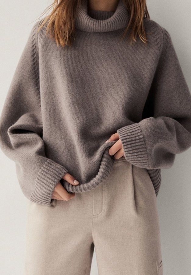 Femme portant un pull col roulé ample taupe à manches longues et un pantalon taille haute beige clair, tenant l'ourlet du pull.