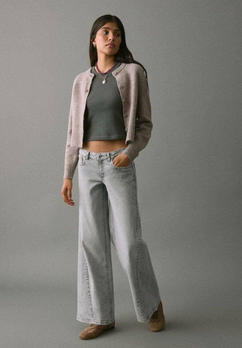 Cardigan corto grigio chiaro su una maglietta attillata grigio scuro, abbinato a jeans in denim azzurro chiaro a gamba larga. Tessuti morbidi, design minimalista, vestibilità casual.