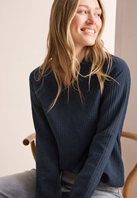 Navyblaues Langarm-Oberteil mit strukturiertem Stoff und geknöpftem Henley-Ausschnitt. Das Model sitzt auf einem Holzstuhl und trägt hellgraue Hosen.